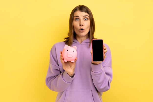 Mujer sorprendida con la boca abierta que muestra el teléfono inteligente con pantalla vacía para promoción y alcancía rosa