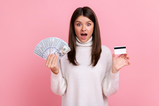 Mujer sorprendida con cabello oscuro sosteniendo un ventilador de billetes en dólares y tarjeta de crédito, ganando dinero, banca, usando suéter blanco de estilo casual. Disparo de estudio interior aislado sobre fondo rosa.