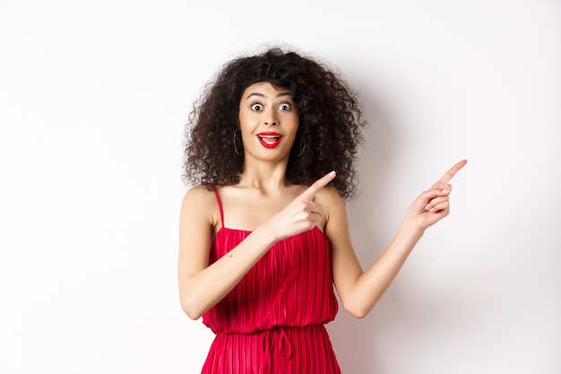 Mujer sorprendida con cabello rizado, maquillaje y vestido rojo, mirando sorprendida y feliz mientras apunta con el dedo directamente al logo, mostrando publicidad, fondo blanco.