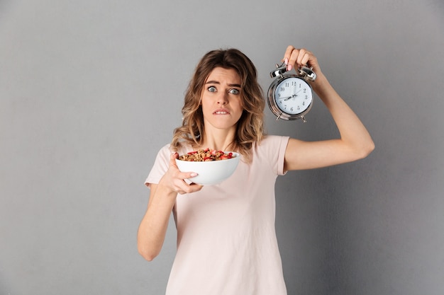 Mujer sorprendida en camiseta con comida sana en plato y reloj despertador mientras se muerde el labio y sobre gris