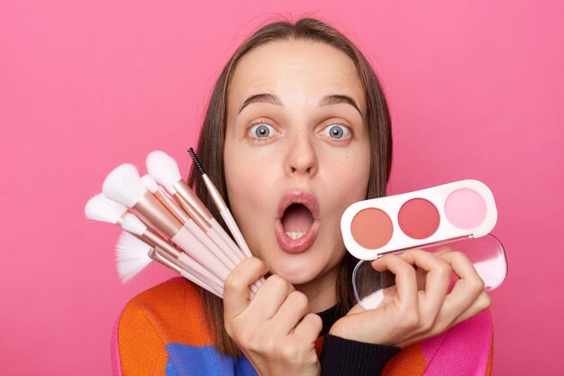 Mujer sorprendida conmocionada usando suéter sosteniendo cepillos y posando en polvo aislada en un fondo rosado mirando con grandes ojos asombrados con nuevos productos cosméticos