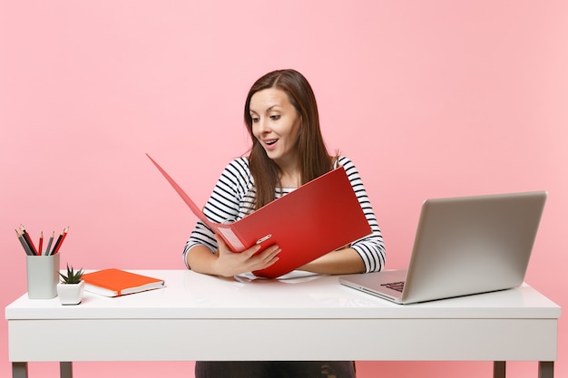 Mujer sorprendida mirando la carpeta roja con documentos de trabajo en papel trabajando en el proyecto mientras está sentado en la oficina con el portátil