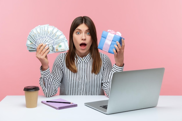 Mujer sorprendida oficinista sosteniendo billetes de dólar y caja de regalo mirando a la cámara con asombro sorprendida con el presente reembolso y bonificaciones Estudio interior aislado en fondo rosa