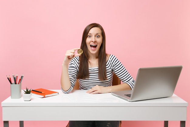 Mujer sorprendida que sostiene bitcoin, moneda de metal de color dorado, moneda futura sentarse a trabajar en un escritorio blanco con un ordenador portátil aislado sobre fondo rosa pastel. Concepto de carrera empresarial de logro. Copie el espacio.