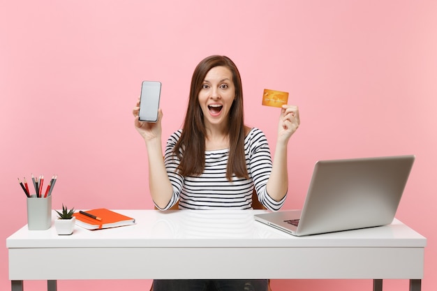 Mujer sorprendida que sostiene el teléfono móvil con la pantalla vacía en blanco, tarjeta de crédito sentarse a trabajar en el escritorio blanco con un portátil pc contemporáneo