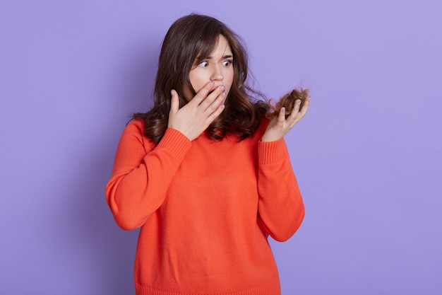 Mujer sorprendida que tiene problemas con la caída del cabello, sostiene el rizo y se cubre la boca con la palma, la dama usa un jersey naranja casual, está frustrada por los problemas de calvicie, aislada sobre una pared azul