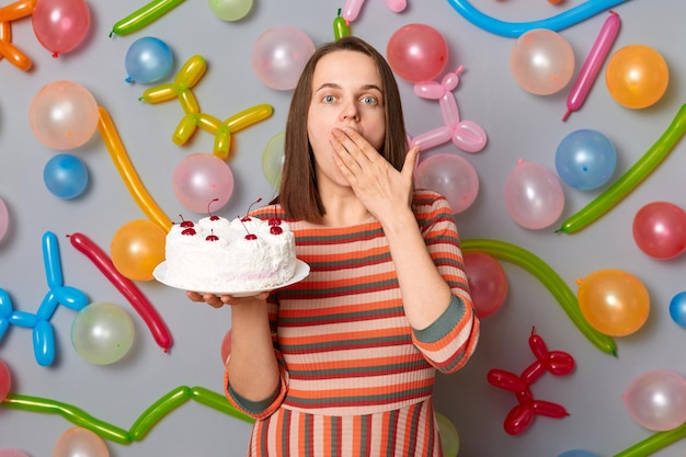 Mujer sorprendida sorprendida con cabello castaño con vestido a rayas sosteniendo pastel cubriendo la boca con las manos ve un regalo de cumpleaños sorprendido de pie contra una pared gris decorada con globos de colores