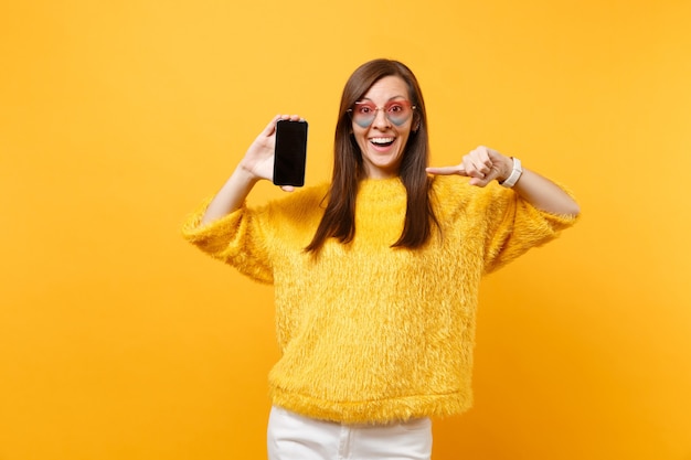 Mujer sorprendida sorprendida en gafas de corazón que señala el dedo índice en el teléfono móvil con la pantalla vacía negra en blanco aislada en fondo amarillo brillante. Personas sinceras emociones, estilo de vida. Área de publicidad.