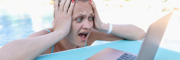 Mujer sorprendida sosteniendo su cabeza y mirando la pantalla del portátil en el lado de la piscina