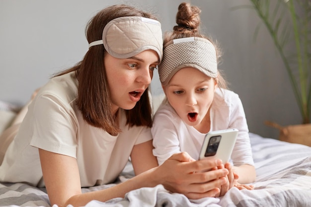 Una mujer sorprendida y su hija con una máscara para dormir acostada en la cama y usando un teléfono móvil mirando la pantalla del dispositivo con la boca abierta revisando las redes sociales