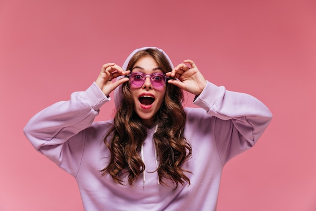 Mujer sorprendida en sudadera con capucha púrpura se quita las gafas de sol brillantes