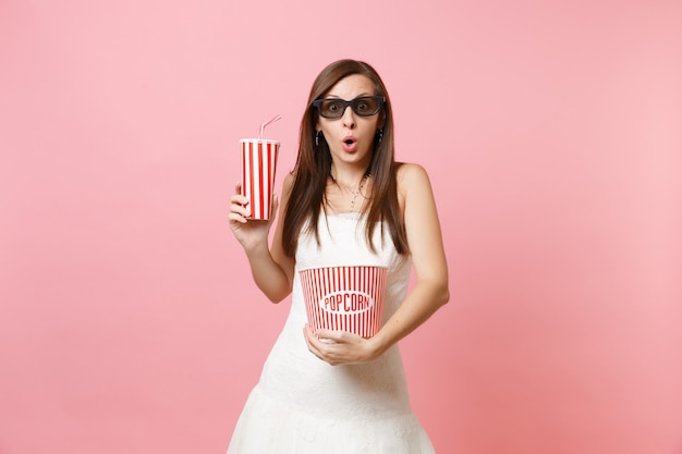 Mujer sorprendida en vestido blanco, gafas 3d viendo una película, sosteniendo un cubo de palomitas de maíz, un vaso de plástico de refresco o cola