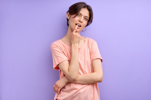 Mujer sospechosa pensando y tocando la boca con el dedo, mire a la cámara, en traje casual, aislado sobre fondo púrpura