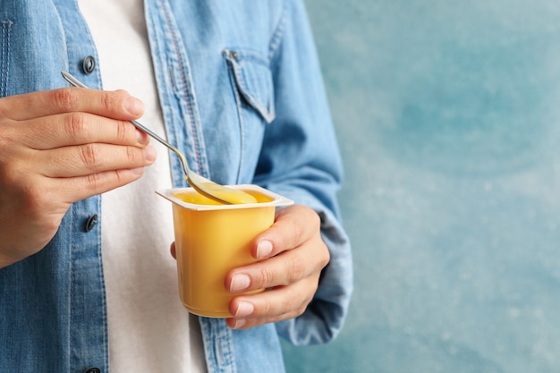 Mujer sostenga un vaso de plástico de yogur de crema agria y una cuchara contra la pared azul