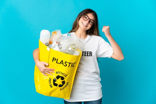 Mujer sosteniendo una bolsa llena de botellas para reciclar aislado en azul celebrando una victoria