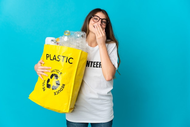 Mujer sosteniendo una bolsa llena de botellas para reciclar aislado en azul feliz y sonriente cubriendo la boca con la mano