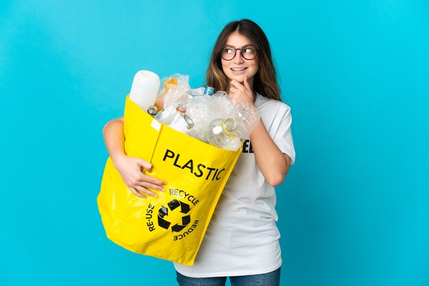 Mujer sosteniendo una bolsa llena de botellas para reciclar aislado en azul mirando hacia el lado y sonriendo