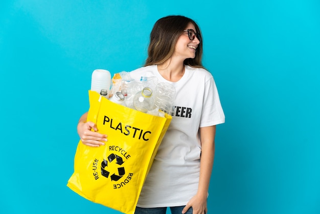 Mujer sosteniendo una bolsa llena de botellas para reciclar aislado en azul riendo en posición lateral