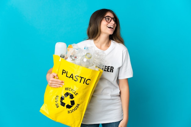 Mujer sosteniendo una bolsa llena de botellas para reciclar aislado en azul riendo