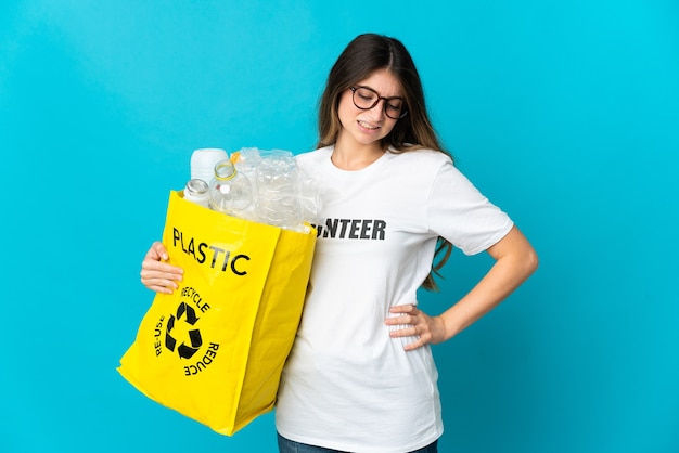 Mujer sosteniendo una bolsa llena de botellas para reciclar aislado en la pared azul que sufre de dolor de espalda por haber hecho un esfuerzo