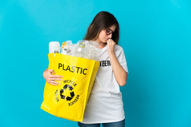 Mujer sosteniendo una bolsa llena de botellas para reciclar aislado en la pared azul que tiene dudas