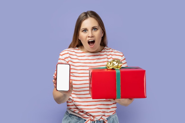 Mujer sosteniendo una caja de regalo y mostrando un teléfono celular con pantalla blanca con espacio de copia para publicidad