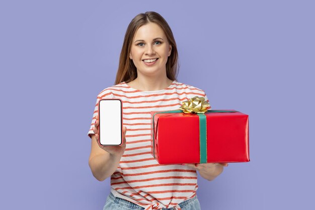 Mujer sosteniendo una caja de regalo roja y mostrando un teléfono inteligente con una pantalla blanca vacía