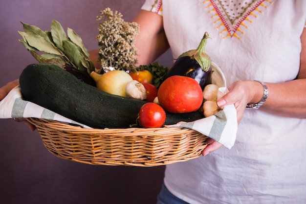 Una mujer sosteniendo una canasta con tomates, calabacines, cebollas, ajo y otros vegetales de temporada