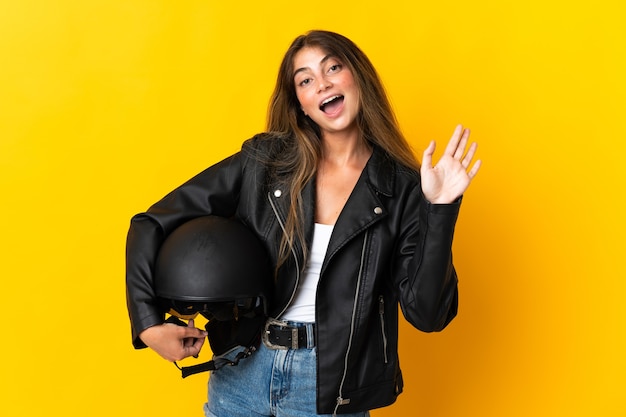 Mujer sosteniendo un casco de motocicleta aislado en amarillo saludando con la mano con expresión feliz