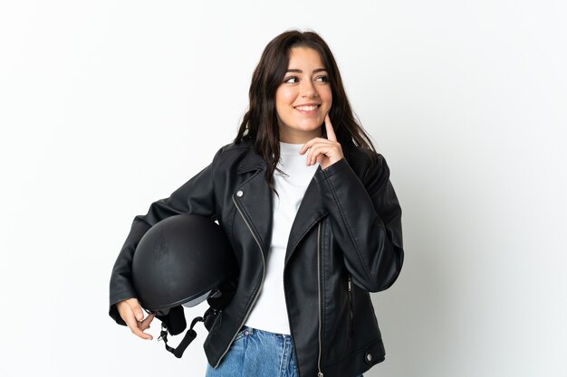 Mujer sosteniendo un casco de motocicleta aislado en la pared blanca pensando en una idea mientras mira hacia arriba