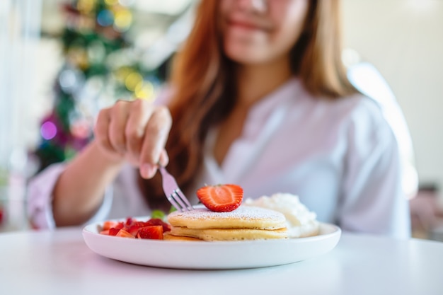 Una mujer sosteniendo y comiendo panqueques con fresas y crema batida con un tenedor