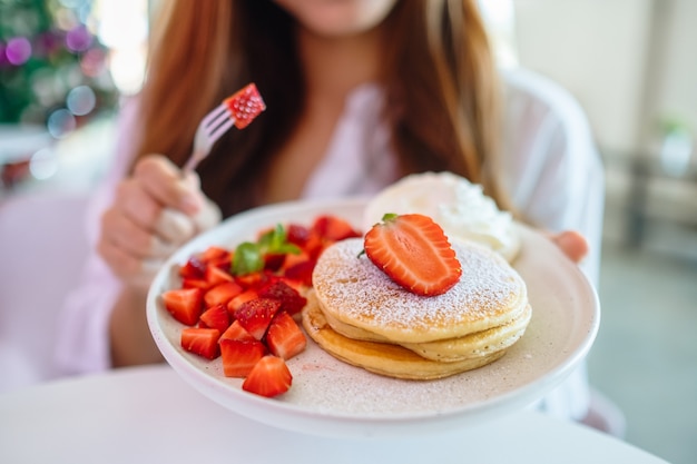 Una mujer sosteniendo y comiendo panqueques con fresas y crema batida con un tenedor