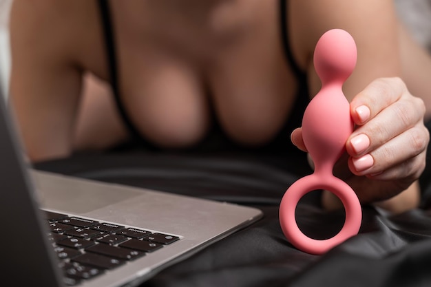 Mujer sosteniendo cuentas anales rosas junto a la computadora portátil mientras está acostada en una sábana negra