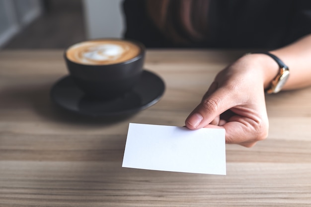 Mujer sosteniendo y dando una tarjeta de negocios vacía a alguien con taza de café en la mesa