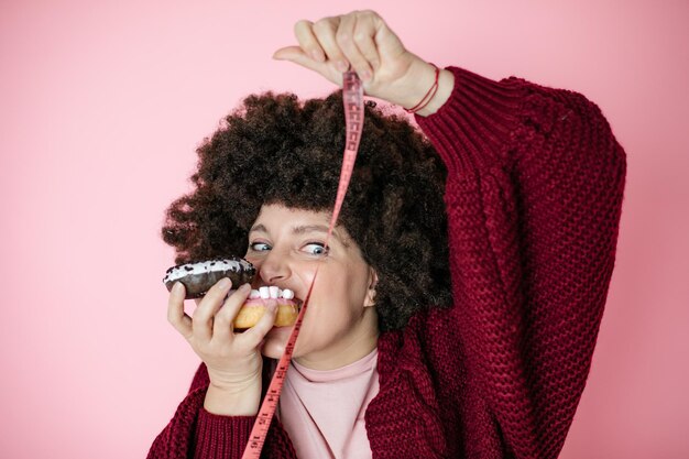 Mujer sosteniendo una dona en la mano y revise su grasa corporal con cinta métrica. foto de humor, diversión, alegría, dieta, concepto de adicción, fondo rosa, peinado afro