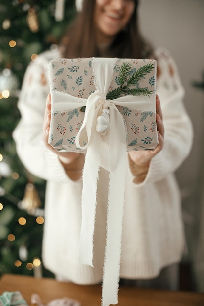 Mujer sosteniendo un elegante regalo de Navidad con cinta y rama de abeto cerca en el fondo de un árbol decorado moderno en la habitación escandinava Feliz Navidad y felices fiestas