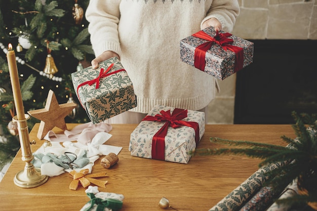 Mujer sosteniendo elegantes regalos de Navidad con cinta roja en una mesa de madera con decoraciones festivas en el fondo de un árbol decorado y una chimenea en una habitación escandinava Feliz Navidad