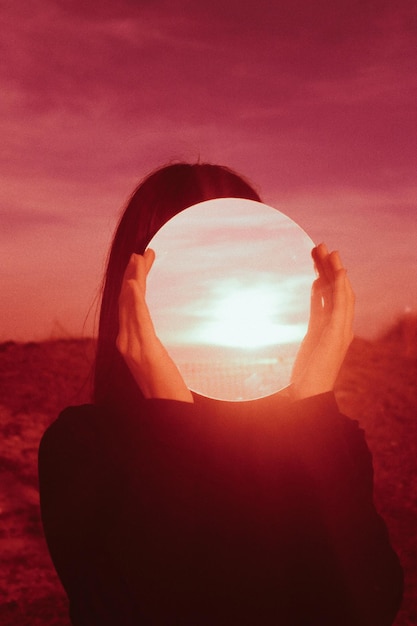 Mujer sosteniendo un espejo con reflejo mientras está de pie contra el cielo durante la puesta de sol