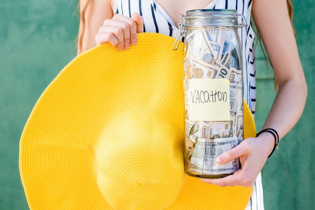 Mujer sosteniendo un frasco lleno de ahorros de dinero para las vacaciones de verano con sombrero amarillo sobre fondo verde