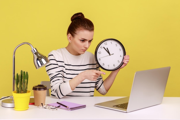 Mujer sosteniendo la gestión del tiempo del reloj de pared grande apuntando a la pantalla del portátil con expresión seria