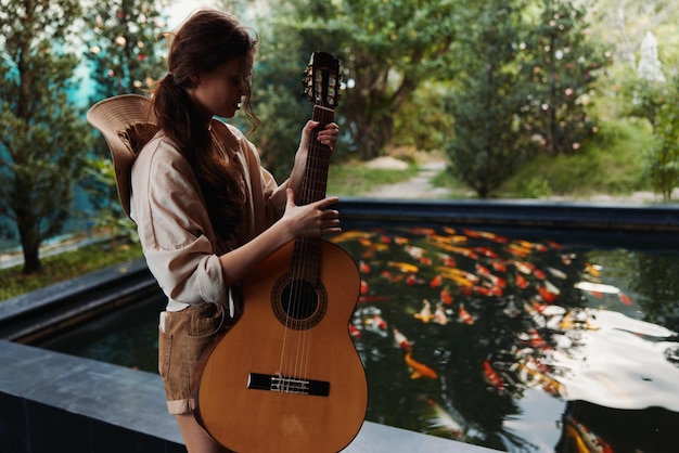 Una mujer sosteniendo una guitarra acústica junto a un estanque con peces dorados en el fondo