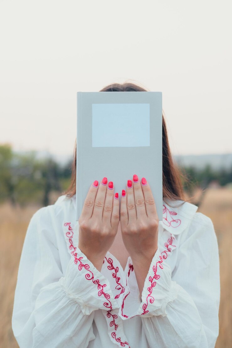 Foto mujer sosteniendo un libro con el rostro cubierto en un prado al atardecer