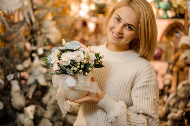 Mujer sosteniendo una maceta con flores blancas decoradas con hojas verdes y ramas de abeto