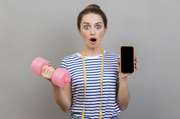 Mujer sosteniendo mancuerna rosa y teléfono celular con pantalla en blanco para texto promocional