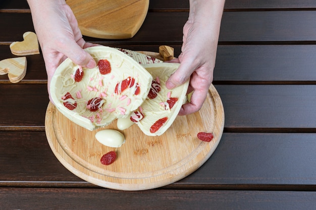 Mujer sosteniendo la mitad de un huevo de Pascua roto de chocolate blanco con fresas confitadas sobre una tabla de madera junto a corazones