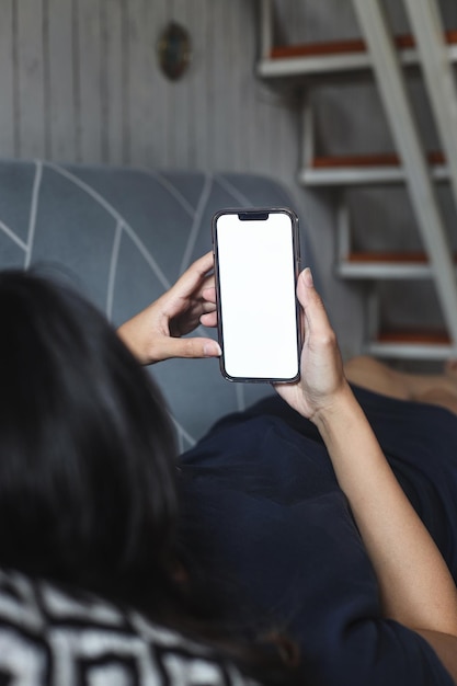 Mujer sosteniendo una pantalla en blanco de teléfono inteligente mientras está acostado en el sofá.