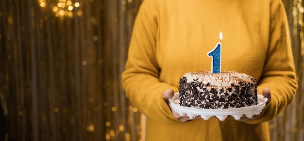 Mujer sosteniendo pastel de cumpleaños con velas encendidas número 1 contra un fondo decorado de cerca Celebra las vacaciones en casa
