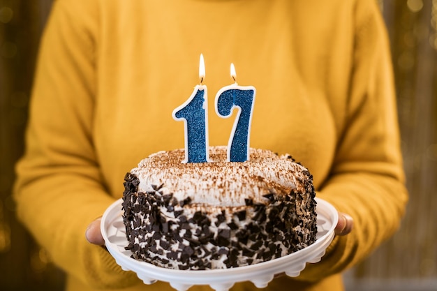 Mujer sosteniendo pastel de cumpleaños con velas encendidas número 17 contra un fondo decorado de cerca Celebra el cumpleaños en casa