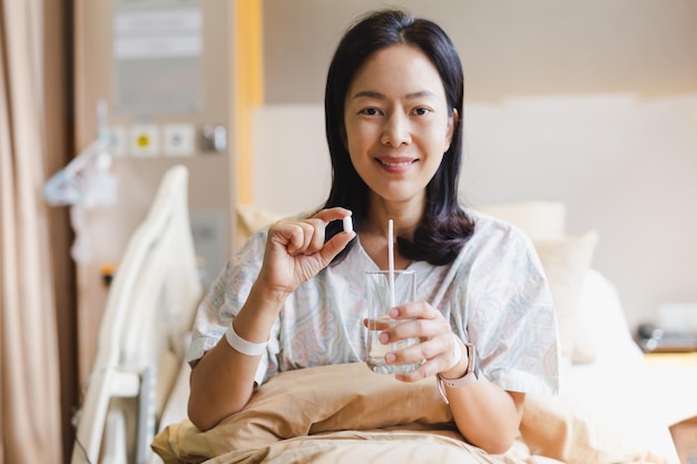 Mujer sosteniendo pastillas blancas y un vaso de agua mientras está sentada en una cama en el hospital