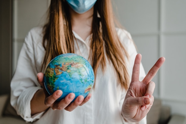 Mujer sosteniendo el planeta tierra en casa con máscara que muestra gesto de paz